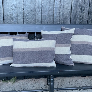 May include: Five decorative throw pillows with a striped design in shades of gray and beige. The pillows are arranged on a black bench, creating a cozy and inviting aesthetic. The pillows are rectangular and have a textured appearance.