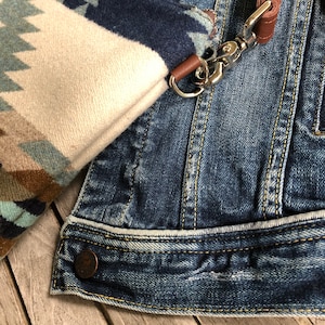 May include: A close-up of a denim jacket with a brown leather strap and a silver clasp. The jacket has a white and blue patterned fabric attached to it.