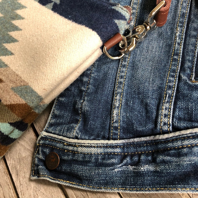 May include: A close-up of a denim jacket with a brown leather strap and a silver clasp. The jacket has a white and blue patterned fabric attached to it.