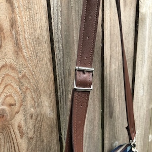 May include: Close-up of a brown leather strap with a silver buckle. The strap is attached to a bag, but the bag is not visible in the image.
