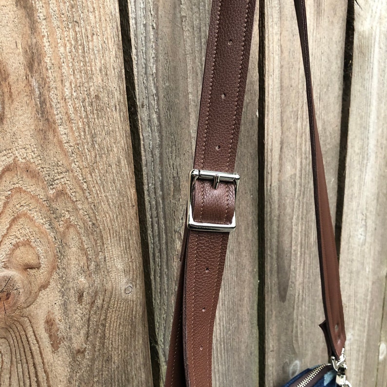May include: Close-up of a brown leather strap with a silver buckle. The strap is attached to a bag, but the bag is not visible in the image.
