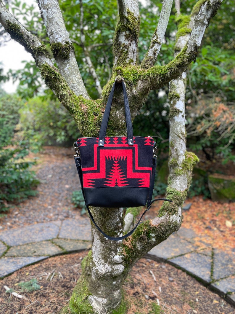 K&ouml;nnte beinhalten: Eine schwarze und rote Tragetasche mit geometrischem Muster h&auml;ngt an einem Baumzweig. Die Tasche hat einen langen Riemen und einen Rei&szlig;verschluss oben.