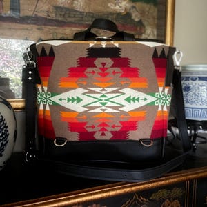 Puede incluir: Una mochila marr&oacute;n y negra con un patr&oacute;n geom&eacute;trico colorido. La mochila tiene una correa de cuero negra y un asa superior.