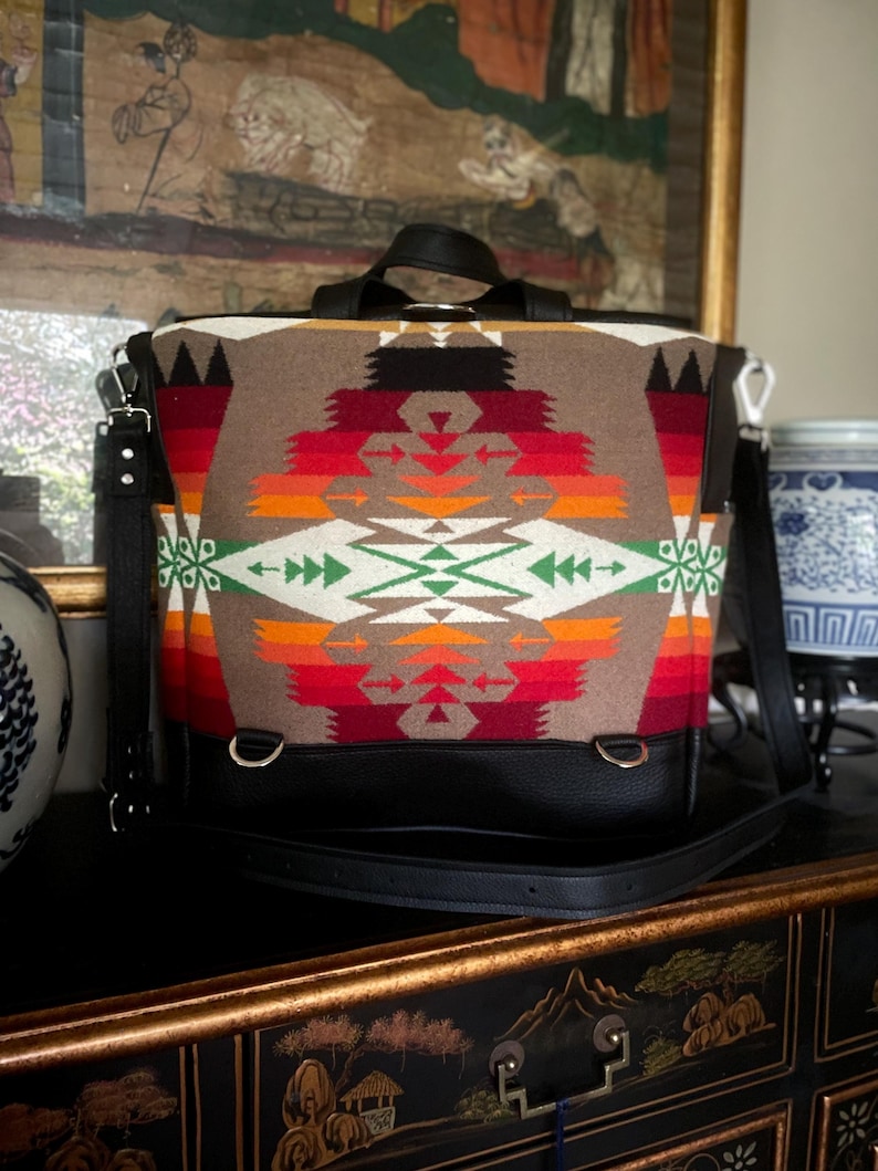 Puede incluir: Una mochila marr&oacute;n y negra con un patr&oacute;n geom&eacute;trico colorido. La mochila tiene una correa de cuero negra y un asa superior.