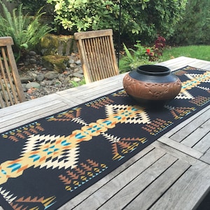 May include: A black and white geometric table runner with orange, yellow, and blue accents. The runner is laid out on a wooden table with a brown pottery bowl in the center.