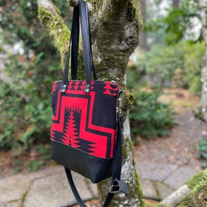 K&ouml;nnte beinhalten: Eine schwarze und rote Tragetasche mit geometrischem Muster. Die Tasche hat einen schwarzen Lederriemen und einen Rei&szlig;verschluss.