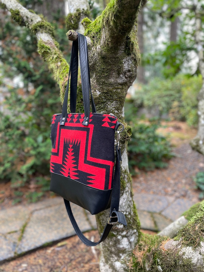 K&ouml;nnte beinhalten: Eine schwarze und rote Tragetasche mit geometrischem Muster. Die Tasche hat einen schwarzen Lederriemen und einen Rei&szlig;verschluss.