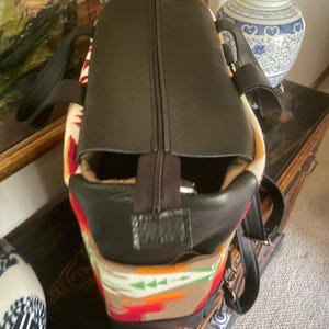 Puede incluir: Una bolsa de viaje de cuero marr&oacute;n con cierre de cremallera y un patr&oacute;n tejido colorido en la parte inferior. La bolsa tiene dos asas y una correa para el hombro.