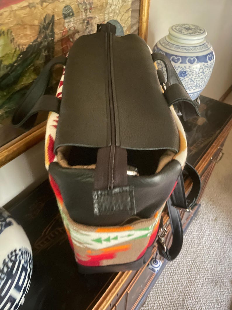 Puede incluir: Una bolsa de viaje de cuero marr&oacute;n con cierre de cremallera y un patr&oacute;n tejido colorido en la parte inferior. La bolsa tiene dos asas y una correa para el hombro.