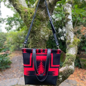K&ouml;nnte beinhalten: Eine schwarze und rote gemusterte Tragetasche mit schwarzen Lederriemen und einem Rei&szlig;verschluss. Die Tasche h&auml;ngt an einem Baumzweig.
