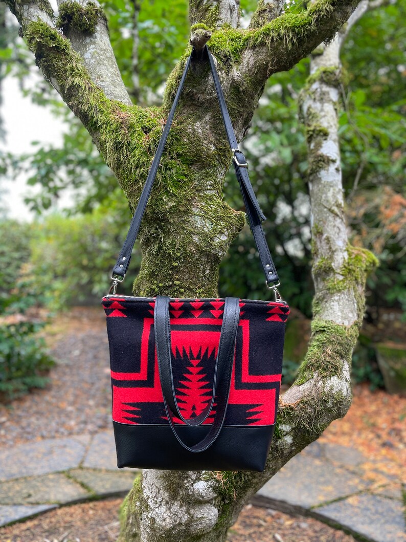 K&ouml;nnte beinhalten: Eine schwarze und rote gemusterte Tragetasche mit schwarzen Lederriemen und einem Rei&szlig;verschluss. Die Tasche h&auml;ngt an einem Baumzweig.