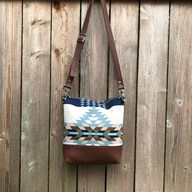 May include: A brown leather crossbody bag with a geometric patterned fabric front. The pattern is in shades of blue, white, and brown.