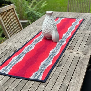 May include: Red and blue patterned table runner with a white and gray design. The runner is on a wooden table with two white ceramic vases.