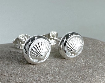May include: A pair of silver stud earrings with a raised seashell design. The earrings are on a gray stone background.
