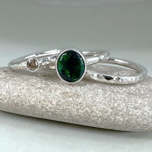 May include: Three silver rings with textured bands. One ring has a brown gemstone, another a large oval green gemstone, and the third is a plain band. The rings are displayed on a grey stone surface.