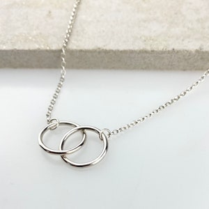 Könnte beinhalten: Eine silberne Kettenkette mit einem Doppelkreis-Anhänger. Die Kreise sind ineinander verschlungen und die Kette ist zart.
