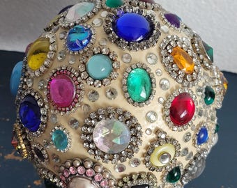 May include: A spherical ornament in cream, adorned with an array of colourful gemstones and rhinestones. The gems are in shades of blue, green, yellow, pink, and red, set in silver-toned metal. The overall effect is ornate and decorative.