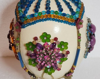 May include: A white ostrich egg decorated with colourful beads, rhinestones, and jewels. The egg is mounted on a gold metal stand with three legs shaped like stylised bird feet.