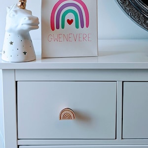 Peut inclure: Une toile blanche avec un arc-en-ciel peint en rose, violet et vert. L'arc-en-ciel a un petit cœur rouge au centre. Le nom "GWENEVERE" est écrit en rouge sous l'arc-en-ciel.