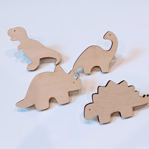 May include: Four wooden dinosaur shapes, including a Tyrannosaurus Rex, a Triceratops, a Stegosaurus, and a Brachiosaurus. The dinosaurs are all cut out of light brown wood and are about 10 cm tall.