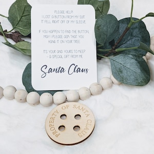 May include: A white card with a handwritten note that reads "PLEASE HELP! I LOST A BUTTON FROM MY SUIT IT FELL RIGHT OFF OF MY SLEEVE IF YOU HAPPEN TO FIND THE BUTTON, MAY I PLEASE ASK THAT YOU HANG IT ON YOUR TREE ITS YOUR AND YOURS TO KEEP - A SPECIAL GIFT FROM ME Santa Claus" and a wooden button with the text "PROPERTY OF SANTA CLAUS" on it.