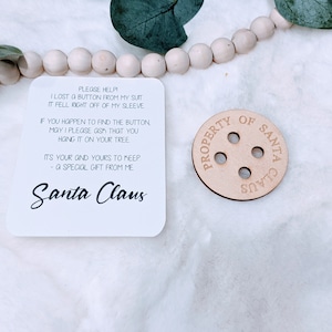 May include: A white card with black text that reads "PLEASE HELP I LOST A BUTTON FROM MY SUIT IT FELL RIGHT OFF OF MY SLEEVE IF YOU HAPPEN TO FIND THE BUTTON MAY I PLEASE ASK THAT YOU HANG IT ON YOUR TREE IT'S YOUR AND YOURS TO KEEP - A SPECIAL GIFT FROM ME Santa Claus" and a wooden button with the text "PROPERTY OF SANTA CLAUS" on it.