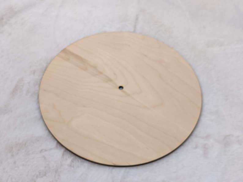 May include: A round, unfinished wooden craft blank with a natural wood grain. The blank has a small hole drilled near the center. This versatile craft supply is ideal for various DIY projects, including signs and home decor.