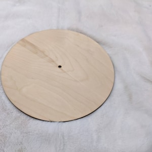 May include: A round, unfinished wooden craft blank. The light-colored wood has visible grain and a small hole in the center. The edge is dark. This blank is suitable for various DIY projects.