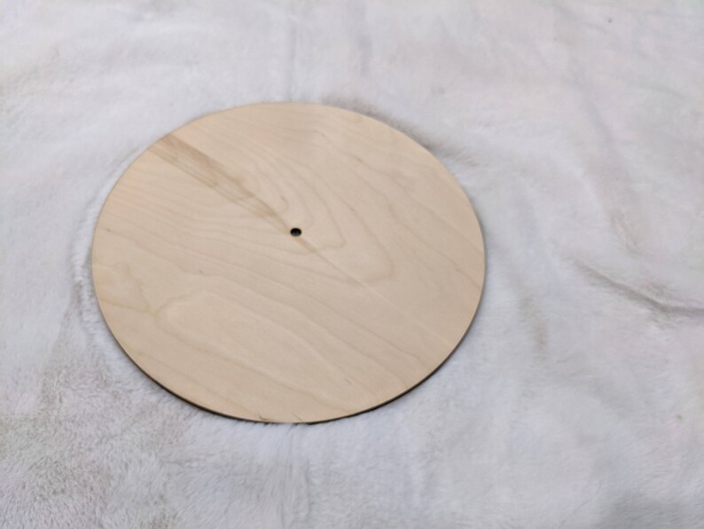 May include: A round, unfinished wooden craft blank. The light-colored wood has visible grain and a small hole in the center. The edge is dark. This blank is suitable for various DIY projects.