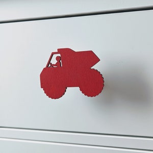 May include: A red wooden truck-shaped drawer pull with a driver silhouette. The pull has two large, textured wheels and is mounted on a white drawer front. The design is simple and child-friendly.