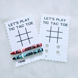 May include: Two white cards with scalloped edges, each with the text "Let's Play Tic Tac Toe" and a tic-tac-toe grid. Two crayons, one red and one blue, are placed on top of the cards.
