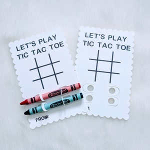 May include: Two white cards with scalloped edges, each with the text "Let's Play Tic Tac Toe" and a tic-tac-toe grid. Two crayons, one red and one blue, are placed on top of the cards.