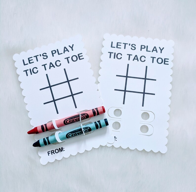 May include: Two white cards with scalloped edges, each with the text "Let's Play Tic Tac Toe" and a tic-tac-toe grid. Two crayons, one red and one blue, are placed on top of the cards.