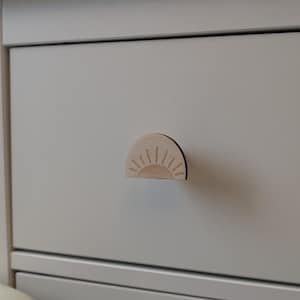 May include: A light-colored wooden drawer pull with a sunburst design. The half-circle pull is attached to a light gray drawer. The sunburst design features a series of thin lines radiating from a curved base.