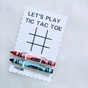 May include: A white card with the text "Let's Play Tic Tac Toe" and a tic-tac-toe grid. Two crayons, one red and one blue, are attached to the card with white paper clips. The card also has the text "From:" at the bottom.