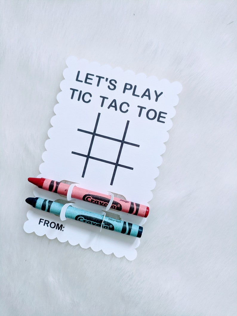 May include: A white card with the text "Let's Play Tic Tac Toe" and a tic-tac-toe grid. Two crayons, one red and one blue, are attached to the card with white paper clips. The card also has the text "From:" at the bottom.