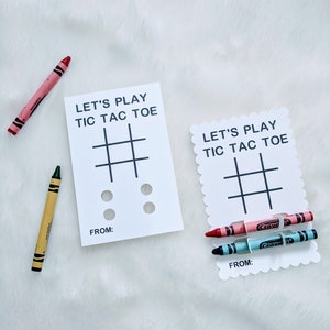 May include: Two white cards with the text "Let's Play Tic Tac Toe" and a tic-tac-toe grid. The cards have a space for a name at the bottom. One card has two red crayons and one blue crayon, the other card has two black circles.