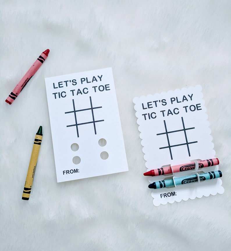 May include: Two white cards with the text "Let's Play Tic Tac Toe" and a tic-tac-toe grid. The cards have a space for a name at the bottom. One card has two red crayons and one blue crayon, the other card has two black circles.