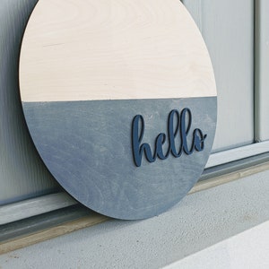 May include: A round wooden sign with a light wood top half and a dark grey bottom half. The word "hello" is written in black lettering on the bottom half of the sign.