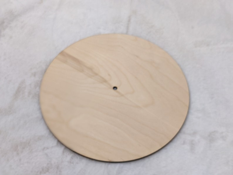 May include: A round, unfinished wooden disc with a small hole in the center. The light-colored wood has a natural grain pattern. This blank wooden circle is suitable for various craft projects.