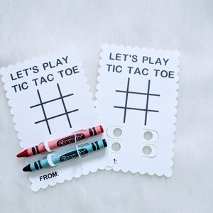 May include: Two white cards with scalloped edges, each with the text "Let's Play Tic Tac Toe" and a tic-tac-toe grid. One card has two circles with a number "1" below them. Two crayons, one red and one blue, are placed on the cards.