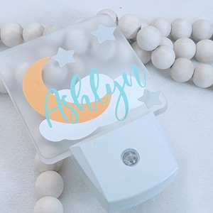 May include: A white night light with a clear plastic cover featuring a crescent moon, stars, and the name "Ashlyn" in teal lettering. The moon is orange and the stars are white.