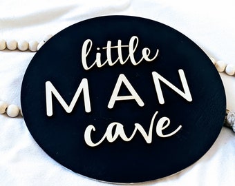 May include: Black round wooden sign with white lettering that reads "little MAN cave".
