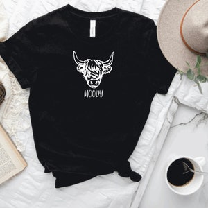 May include: Black t-shirt featuring a white graphic of a Highland cow's head with the word "MOODY" underneath.