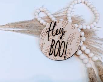 May include: A round wooden sign with a ghost pattern and the text "Hey BOO!" in black lettering. The sign is surrounded by white beads and dried pampas grass.