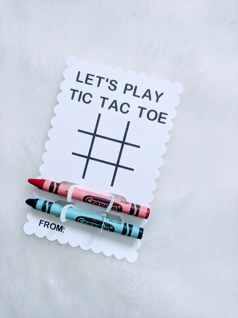 May include: A white card with the words "Let's Play Tic Tac Toe" and a tic-tac-toe grid. Two crayons, one red and one blue, are attached to the card with white paper clips. The card also says "From:" at the bottom.