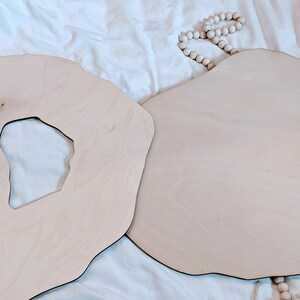 May include: Two irregularly shaped, light-colored wooden boards with a natural wood grain. One board has a hole cut out of the center. The boards are on a white surface, with a strand of wooden beads.