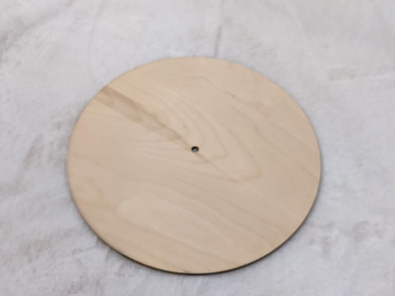 May include: A round, unfinished wooden disc with a small hole in the center. The light-colored wood has a natural grain pattern. This blank wooden circle is suitable for various craft projects.