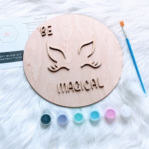 May include: A DIY wood kit for painting a round wooden sign with the words "BE MAGICAL" and two stylized eyes. The kit includes five small paint pots in shades of blue, green, purple, black, and pink, and a paintbrush.