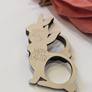 May include: Two wooden bunny napkin rings with the text "Happy Easter" engraved on them. The rings are light brown and have a natural wood finish.